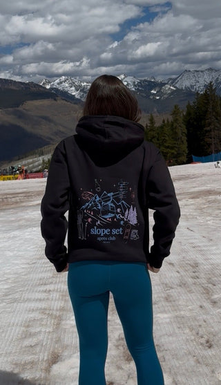 Person wearing a black hoodie and blue pants standing on a snow-covered slope with mountains in the background.