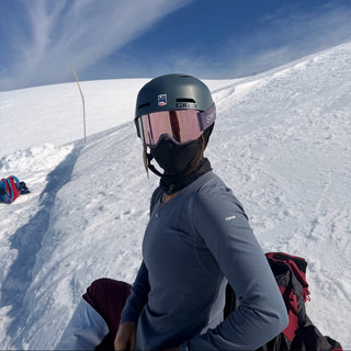 A person wearing a dark gray ski helmet, pink-tinted ski goggles, a black face mask, and a slate blue long-sleeve athletic shirt, sitting on a snowy mountain slope under a bright blue sky.
