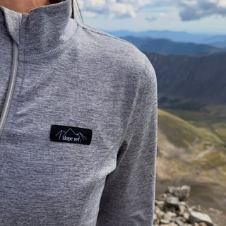 A close-up of a person wearing a heather gray quarter-zip pullover with a black chest patch logo that reads 'slope jet', set against a blurred mountain landscape background.