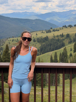 A woman wearing a light blue tank top and matching shorts stands on a wooden deck railing with a scenic mountain valley landscape in the background.